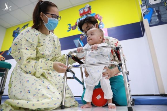 Trúc Nhi lần đầu tiên tập đứng bằng khung đỡ, mẹ bé hạnh phúc động viên con gái. Ảnh: Hữu Khoa.